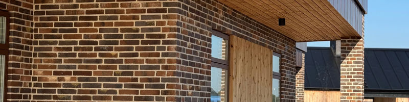 Overdækket terrasse ved moderne nybyggeri med rødbrun murstensfacade, træbeklædt loft, store vinduespartier og træterrasse – stilren inspiration til husdrømme og drømmehuset.