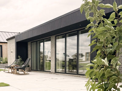 Udvendigt terrassemiljø fra eurodan-huse med store mørke vinduespartier, skydedøre, sort facadebeklædning, murstensdetaljer og loungestole på flisebelagt terrasse – inspiration til nybyggeri, husbyggeri og moderne boliger med tæt kontakt til haven.
