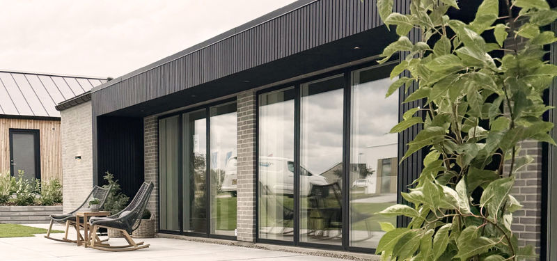 Udvendigt terrassemiljø fra eurodan-huse med store mørke vinduespartier, skydedøre, sort facadebeklædning, murstensdetaljer og loungestole på flisebelagt terrasse – inspiration til nybyggeri, husbyggeri og moderne boliger med tæt kontakt til haven.