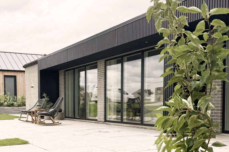 Udvendigt terrassemiljø fra eurodan-huse med store mørke vinduespartier, skydedøre, sort facadebeklædning, murstensdetaljer og loungestole på flisebelagt terrasse – inspiration til nybyggeri, husbyggeri og moderne boliger med tæt kontakt til haven.