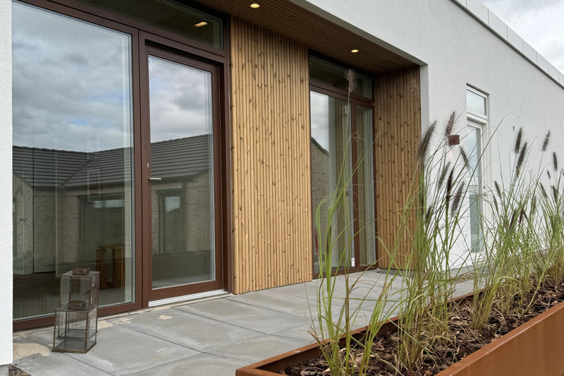 Facadeparti fra eurodan-huse med store brune vinduer, bred terrassedør, lodret træbeklædning, overdækket terrasse og plantekasse foran huset – inspiration til vinduer og døre i moderne nybyggeri og husbyggeri.
