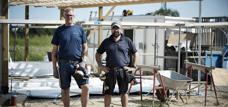To håndværkere står på byggepladsen foran træ- og tagkonstruktionen til et nyt hus under opførelse.
