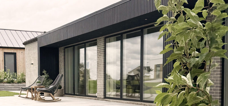 Moderne husfacade i nybyggeri med mørk træbeklædning, store gulv-til-loft-vinduer og flisebelagt terrasse med loungestole.