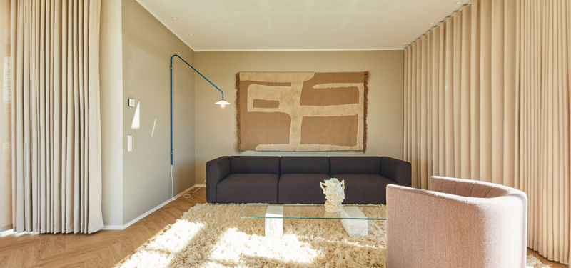 Stue fra eurodan-huse med mørk lav sofa, rosa rund loungestol, glasbord, lang gardinvæg, blød langluvet tæppe, blå væglampe og stort tekstilkunstværk over sofaen – inspiration til nybyggeri, husbyggeri og personlige husdrømme.