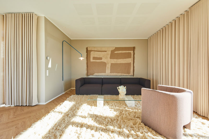 Stue fra eurodan-huse med mørk lav sofa, rosa rund loungestol, glasbord, lang gardinvæg, blød langluvet tæppe, blå væglampe og stort tekstilkunstværk over sofaen – inspiration til nybyggeri, husbyggeri og personlige husdrømme.