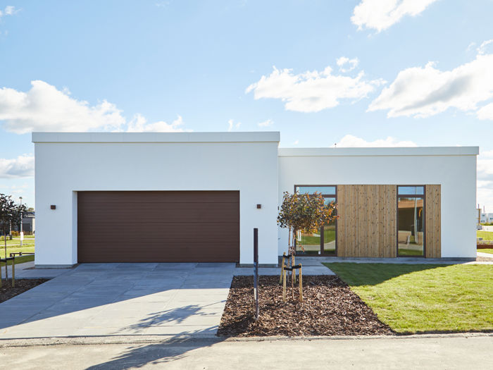 Minimalistisk garage integreret i hvid facade med bred mørk garageport, flad taglinje, træbeklædt indgangsparti og store vinduesfelter – inspiration til moderne garage, nybyggeri og gennemtænkt husbyggeri fra eurodan-huse.