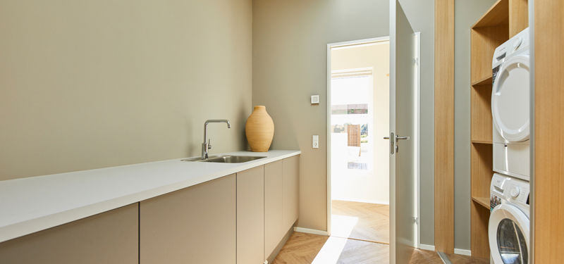 Bryggers fra eurodan-huse med lang hvid bordplade, integreret vask, beige underskabe, keramisk vase på bordet og åben dør mod resten af boligen samt vaskesøjle i træskab ved siden af – inspiration til funktionelt nybyggeri, praktisk husbyggeri og gennemtænkte husdrømme.