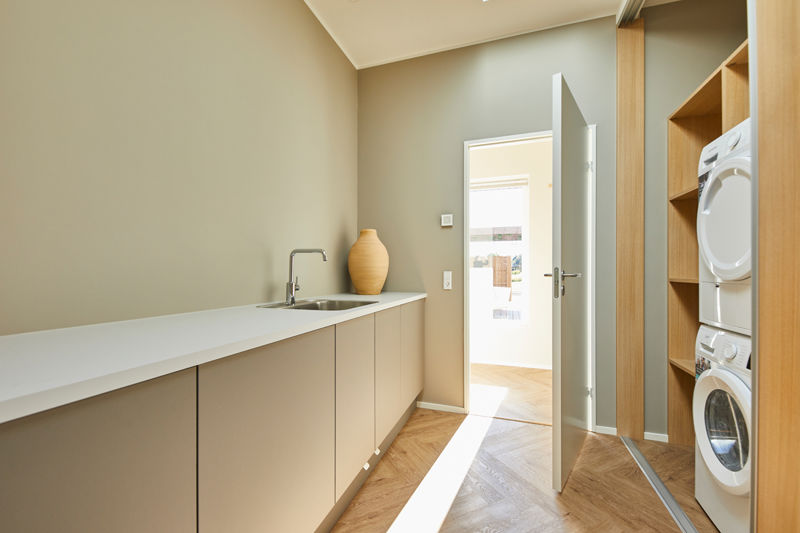 Bryggers fra eurodan-huse med lang hvid bordplade, integreret vask, beige underskabe, keramisk vase på bordet og åben dør mod resten af boligen samt vaskesøjle i træskab ved siden af – inspiration til funktionelt nybyggeri, praktisk husbyggeri og gennemtænkte husdrømme.