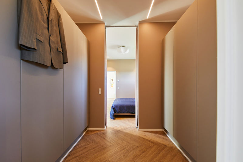 Smal walk-in-gang fra eurodan-huse med høje skabsvægge på begge sider, brune vægge, integrerede lysstriber i loftet, jakker på væggen og direkte kig ind til soveværelse – inspiration til eksklusiv garderobeløsning i nybyggeri og gennemtænkte husdrømme.