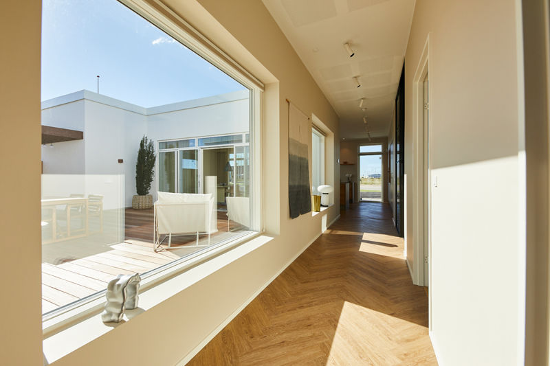 Lang gang fra eurodan-huse med stort fast vindue mod terrasse, indvendige vinduespartier langs væggen, ovenlys i enden og lyst trægulv – inspiration til nybyggeri, husbyggeri og boliger med gennemgående dagslys.