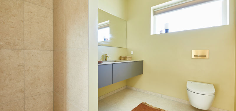 Lyst badeværelse fra eurodan-huse med væghængt toilet, højtsiddende vindue, gråt svævende badmøbel, bredt spejl, lyse fliser omkring bruseområdet og varme beige vægge – inspiration til funktionelt nybyggeri, husbyggeri og enkle husdrømme.