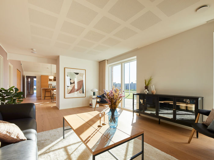 Stue fra eurodan-huse med mørk sofa, lyst sofabord med glasplade, sort vitrineskab, stor plakat på væggen, store vinduer mod terrasse og kig videre til køkken-alrum – inspiration til moderne nybyggeri, husbyggeri og husdrømme.