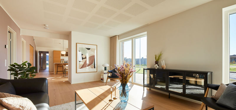 Stue fra eurodan-huse med mørk sofa, lyst sofabord med glasplade, sort vitrineskab, stor plakat på væggen, store vinduer mod terrasse og kig videre til køkken-alrum – inspiration til moderne nybyggeri, husbyggeri og husdrømme.