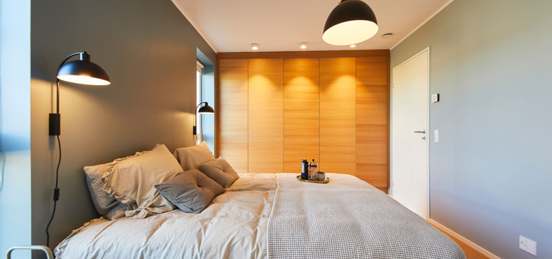 Soveværelse fra eurodan-huse med seng langs grøn væg, beige sengetøj, sorte væglamper, stor skabsvæg i trælook fra gulv til loft, hvid dør og sort pendel over sengen – inspiration til varme materialer i nybyggeri, husbyggeri og personlige husdrømme.