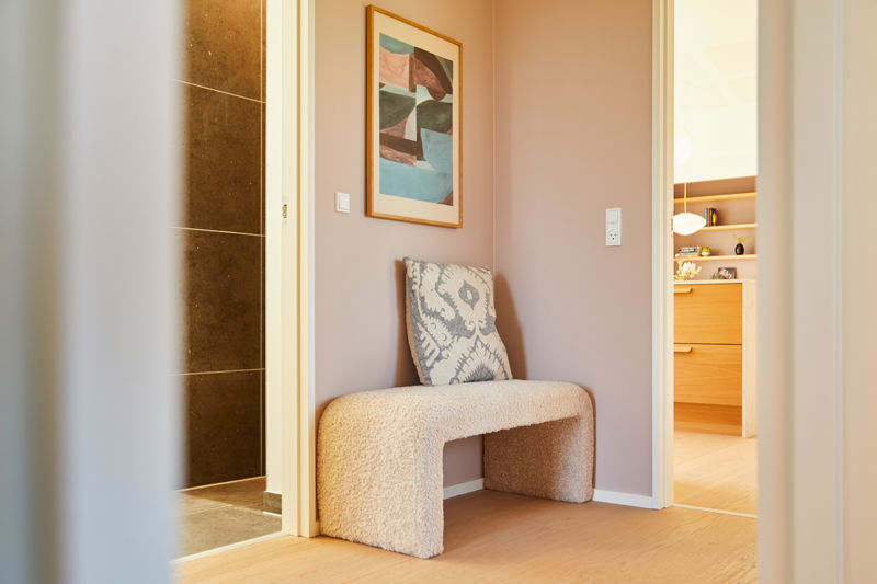 Lille entréniche fra eurodan-huse med rosa væg, polstret bænk, mønstret pude, farverigt vægbillede og kig til både badeværelse og opbevaringsrum – inspiration til personlig entré, nybyggeri og husbyggeri med detaljer.