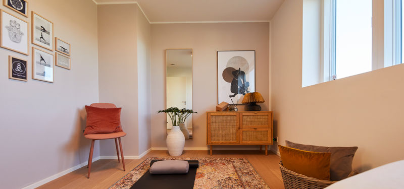Fleksibelt værelse i moderne vinkelhus med yogamåtte, lænestol, kommode, spejl og billedvæg – inspiration til et personligt rum i drømmehuset.