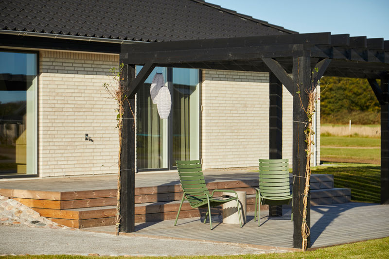 Terrasse fra eurodan-huse med sort pergola, trædæk, grønne havestole, lyse mursten, store vinduespartier og klatreplanter på pergolastolperne – inspiration til hyggeligt uderum, nybyggeri og husdrømme med terrasse.