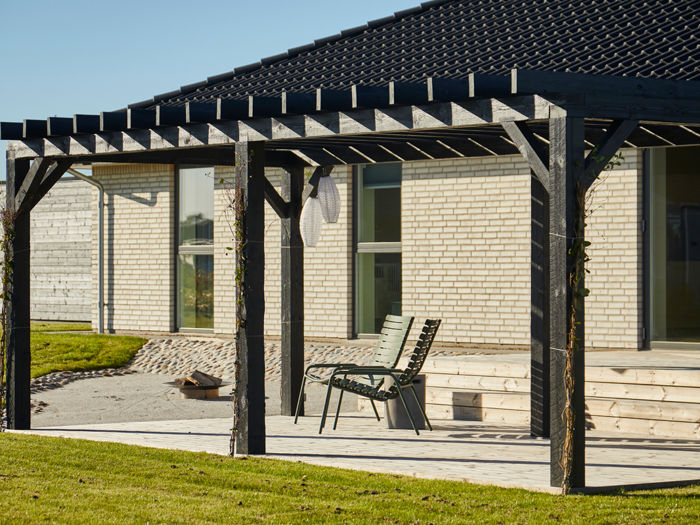 Overdækket terrasse ved længehus i nybyggeri med træpergola, lys murstensfacade og direkte adgang til haven.