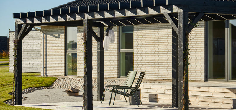 Overdækket terrasse ved længehus i nybyggeri med træpergola, lys murstensfacade og direkte adgang til haven.