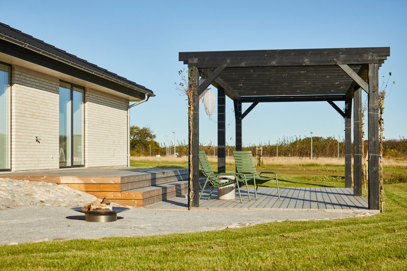 Udemiljø ved moderne længehus i nybyggeri med træterrasse, sort pergola, loungestole og bålsted – inspiration til udeliv ved det nye hus.