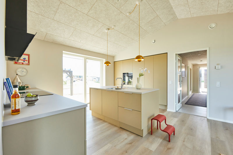 Køkken fra eurodan-huse med beige køkkenø, vask og armatur, lyse skabsvægge med integreret sort ovn, to gule pendler, sort emhætte over kogeplade, lille rød skammel og åben gangzone mod entréen – inspiration til funktionelt nybyggeri, husbyggeri og moderne husdrømme.