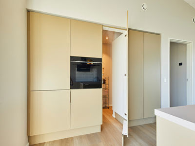 Indbygget skabsvæg i køkken-alrum fra eurodan-huse med lyse beige fronter, integreret sort ovn, åben låge ind til depot/opbevaring, høje skabe fra gulv til loft og lys køkkenø i forgrunden – inspiration til funktionelt nybyggeri, moderne husbyggeri og gennemtænkte husdrømme.