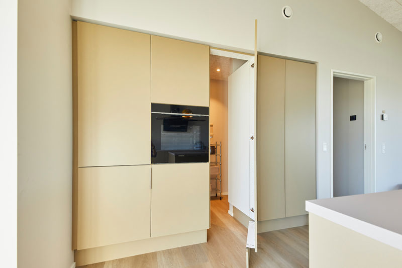 Indbygget skabsvæg i køkken-alrum fra eurodan-huse med lyse beige fronter, integreret sort ovn, åben låge ind til depot/opbevaring, høje skabe fra gulv til loft og lys køkkenø i forgrunden – inspiration til funktionelt nybyggeri, moderne husbyggeri og gennemtænkte husdrømme.