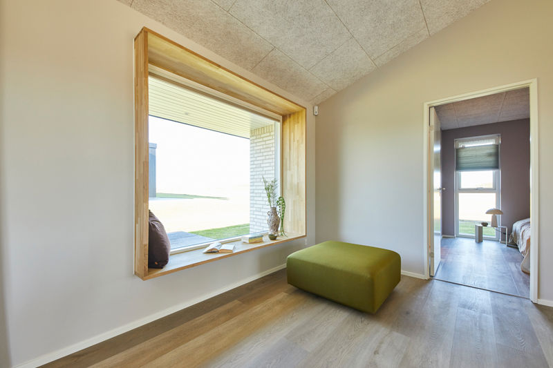 Opholdsrum fra eurodan-huse med store vinduespartier, mørke vinduesrammer, lyse gardiner, siddeplads ved vinduet og åben forbindelse til stueindretning – inspiration til nybyggeri, husbyggeri og moderne vinduesløsninger.