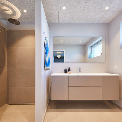 Badeværelse fra eurodan-huse med lyst svævende badmøbel, stor spejlvæg, bruseniche med beige fliser, rainshower, højtsiddende vindue og synligt betonlook loft – inspiration til nybyggeri, husbyggeri og badeværelsesdrømme med rene linjer.