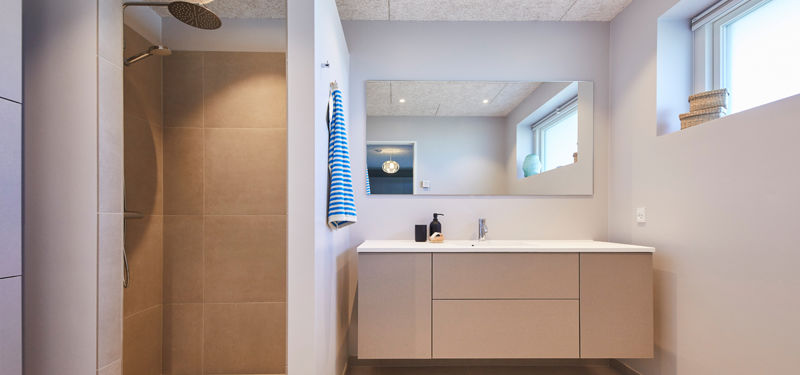 Badeværelse fra eurodan-huse med lyst svævende badmøbel, stor spejlvæg, bruseniche med beige fliser, rainshower, højtsiddende vindue og synligt betonlook loft – inspiration til nybyggeri, husbyggeri og badeværelsesdrømme med rene linjer.