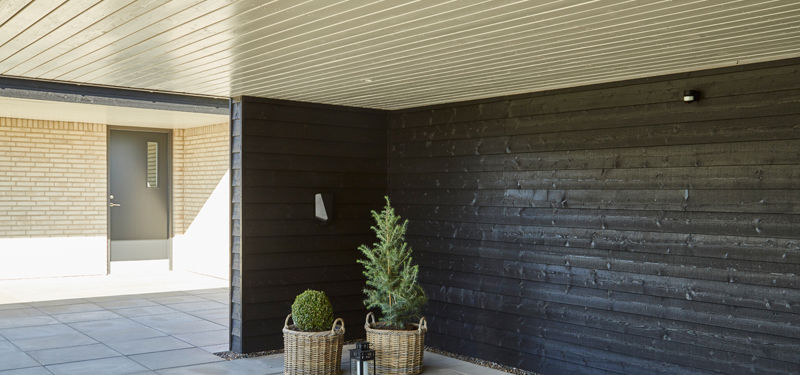 Overdækket carport med sort vandret træbeklædning, lyst træloft, grå fliser, plantekrukker og adgang videre til husets indgang – inspiration til carportindretning, nybyggeri og praktiske løsninger fra eurodan-huse.