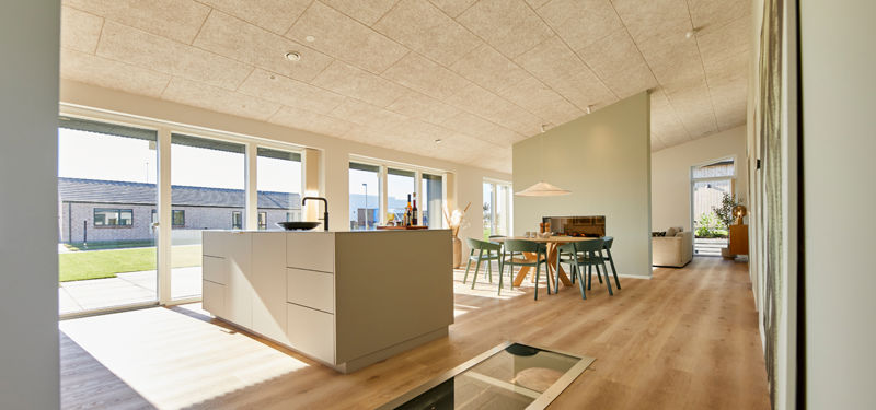 Køkken-alrum i nybyggeri med køkkenø, spiseplads, træbetonloft, trægulv og store vinduespartier med udgang til haven.