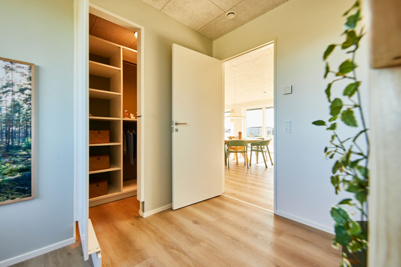 Indgang til walk-in closet fra eurodan-huse med åbne træhylder, kurve, bøjlestang, varm belysning, grønne vægge, åben dør og kig videre til lyst opholdsrum med spiseplads – inspiration til praktisk opbevaring i nybyggeri og personlige husdrømme.