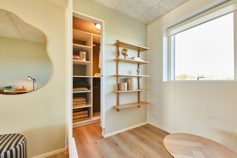Arbejdskrog ved walk-in closet fra eurodan-huse med organisk spejl, lille skriveplads, åbne træhylder, stort vindue, rund bordplade og kig ind til garderobe med hylder og ophæng – inspiration til fleksibel rumudnyttelse i nybygget hus og moderne husbyggeri.