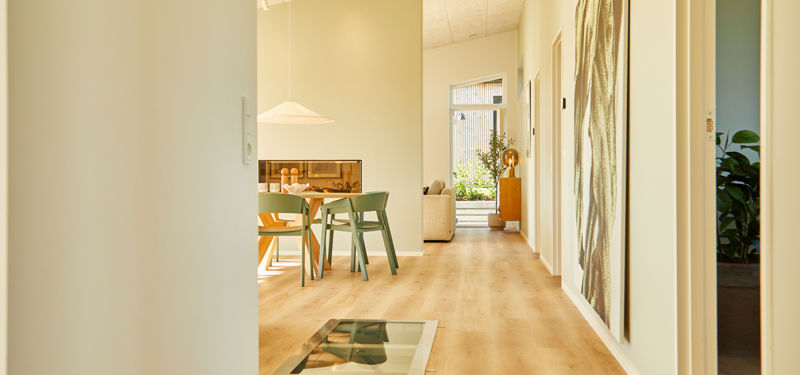 Køkken-alrum i nybyggeri med trægulv, spiseplads, vægkunst og kig gennem huset mod store vinduespartier.