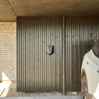 Carport med sort trævæg, vægmonteret ladestander, grå flisebelægning og hvid bil parkeret under mørkt loft – inspiration til carport med elbil-ladning, moderne nybyggeri og husdrømme fra eurodan-huse.