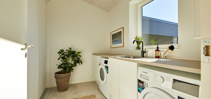 Bryggers fra eurodan-huse med vaskemaskine og tørretumbler under lys bordplade, sort armatur, stort vindue over vasken, hvide skuffer, plante i krukke og lyst gulv – inspiration til nybyggeri med praktisk vaskeplads og moderne husbyggeri.