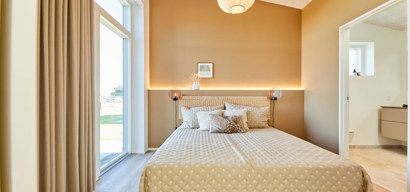 Soveværelse fra eurodan-huse med varm brun væg, vægpanel bag sengen, skjult LED-lys, beige prikket sengetæppe, store vinduer med gardiner og åben dør til badeværelse med lyst badmøbel – inspiration til nybyggeri, husbyggeri og eksklusive husdrømme.