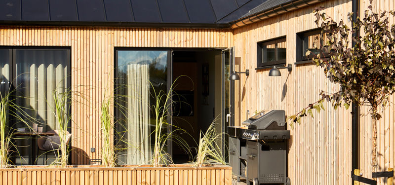 Overdækket terrasse ved nybygget barnhouse med lodret træbeklædning, store glaspartier, loungemøbler og udekøkken til husdrømme med udeliv