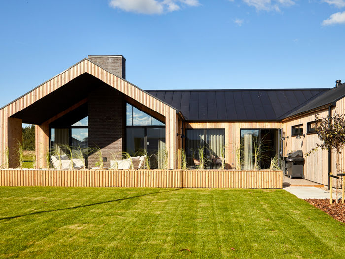 Moderne H-hus med lodret træbeklædning, sort tag, store vinduespartier og overdækket terrasse ud mod en grøn have – inspiration til nybyg og familiens husdrømme.
