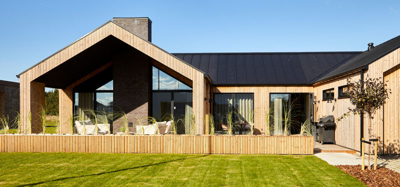 Moderne H-hus med lodret træbeklædning, sort tag, store vinduespartier og overdækket terrasse ud mod en grøn have – inspiration til nybyg og familiens husdrømme.