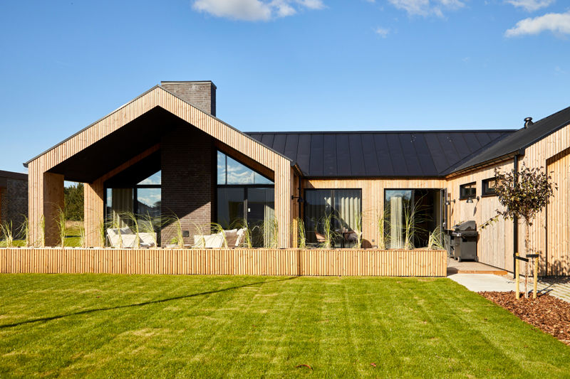 Moderne H-hus med lodret træbeklædning, sort tag, store vinduespartier og overdækket terrasse ud mod en grøn have – inspiration til nybyg og familiens husdrømme.