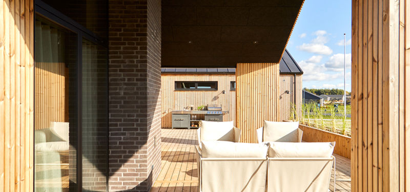 Kig gennem overdækket terrasse i nybygget udstillingshus med loungemøbler, murstenssøjle, udekøkken og varm træbeklædning