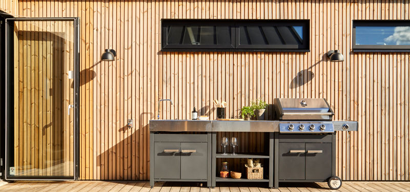 Udekøkken på træterrasse ved nybygget udstillingshus med grill, vask, opbevaring og sort facadebelysning mod lodret træbeklædning