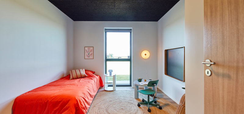Lyst børneværelse med enkeltseng, skriveplads, væghængt skærm, stort vindue og mørkt listeloft – moderne inspiration til nybyg og familiens husdrømme.