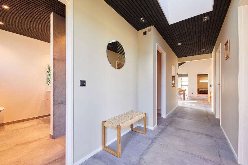 Lang gang fra eurodan-huse med mørkt liste-loft, ovenlys, store grå gulvfliser, rund spejl, flettet bænk og åbning til badeværelse – inspiration til moderne entré, gangareal og nybyggeri med stærke materialevalg.