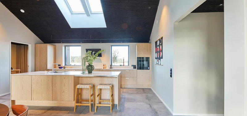 Lyst køkken-alrum med køkken i lyst træ, stor køkkenø, barstole, ovenlysvinduer, mørkt listeloft og gulv i betonlook – moderne inspiration til nybyg og husdrømme.