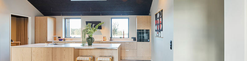 Lyst køkken-alrum med køkken i lyst træ, stor køkkenø, barstole, ovenlysvinduer, mørkt listeloft og gulv i betonlook – moderne inspiration til nybyg og husdrømme.