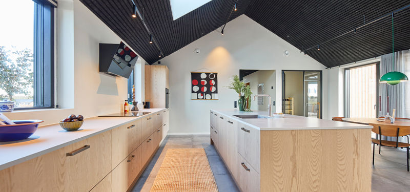 Moderne køkken i lyst træ med stor køkkenø, lange bordflader, ovenlysvindue, mørkt listeloft og gulv i betonlook – stilren inspiration til nybyg og husdrømme.