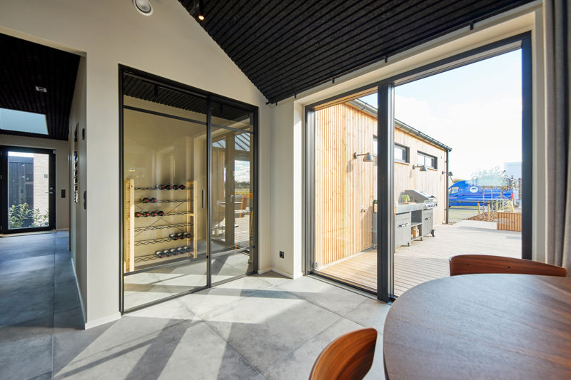 Vinopbevaring i depot fra eurodan-huse med sorte glasvægge, vinreoler langs væggen, store vinduespartier mod terrasse, mørkt akustikloft og spisebord i forgrunden – inspiration til gennemtænkt nybyggeri, moderne husbyggeri og husdrømme med ekstra opbevaringsplads.
